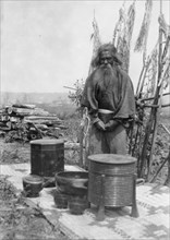 Ainu man standing outdoors on a mat covered with clay containers, 1908. Creator: Arnold Genthe.