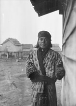 Ainu standing outside by the wall of a wooden hut, 1908. Creator: Arnold Genthe.