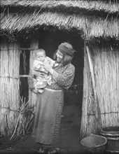 Ainu woman holding a child standing outside a hut, 1908. Creator: Arnold Genthe.
