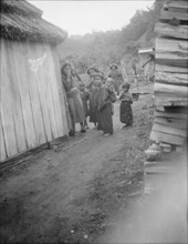 Group of Ainu children standing in a passageway between huts, 1908. Creator: Arnold Genthe.