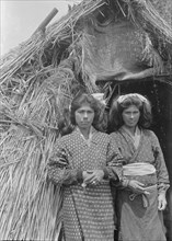Two Ainu women standing at the entrance of a hut, 1908. Creator: Arnold Genthe.