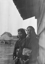 Two Ainu women standing outside by the wall of a wooden hut, 1908. Creator: Arnold Genthe.
