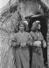 Two Ainu women standing outside the entrance of a hut, 1908. Creator: Arnold Genthe.