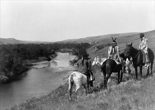 Three Piegan Indians and four horses on hill above river, c1910. Creator: Edward Sheriff Curtis.