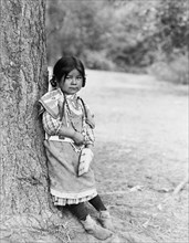 Innocence, an Umatilla girl, full-length portrait, standing by tree, facing slightly right, c1910. Creator: Edward Sheriff Curtis.