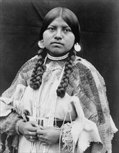 Cayuse woman, half-length portrait, standing, facing front, braids, shell disk earrings..., c1910. Creator: Edward Sheriff Curtis.