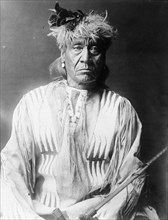 Atsina Indian, Red Whip, half-length portrait, seated, facing front, wearing feather..., c1908. Creator: Edward Sheriff Curtis.