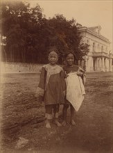 Two barefoot children standing in muddy street in front of the Lindholm's large..., (1899?). Creator: Eleanor Lord Pray.