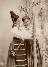 Two young women pose in different national costumes, 1890-1920. Creator: Helene Edlund.