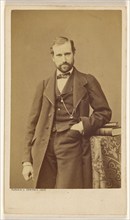 Unidentified well-dressed bearded man standing, elbow on books on a table, 1865-1870. Creator: Bayard & Bertall.