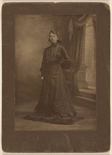 Portrait of a young woman in long black dress, standing, about 1900. Creator: George W Killian.