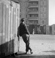 A man leaning on a street corner hoarding looks across the road to where a woman and child pass