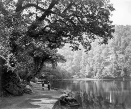 Lake at Naini Tal, India, c1865. Two people stand on the shore beside this picturesque, tree-lined