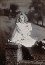 A girl, holding a doll, sitting on a wall, c1890s. Creator: Lock & Whitfield.