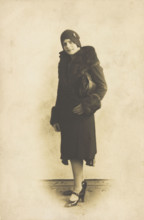 A person poses wearing a long coat with fur trim and a hat with a brooch, [between 1920 and 1929?]. Creator: Unknown.