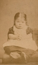 A very young girl, showing signs of mental deficiency, seated with her hands clasped upon her lap. Creator: William Moscrop.
