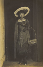 A person wearing a dress, large hat and roller skates, poses holding a basket, (1910-1919?). Creator: Unknown.