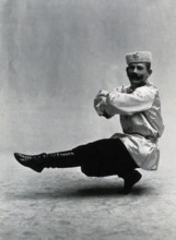 A man, in a satin tunic and fur hat, dancing a "Cossack" measure in a studio setting, 1899-1908. Creator: Unknown.