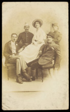 Amateur actors sitting down, surrounding a man in drag seated in the centre, [1910-1919?]. Creator: Unknown.