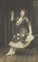A performer in drag, wearing a large flowery dress, poses with a cigarette on stage, c1908-1910. Creator: Unknown.