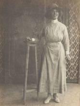 A sailor in drag leans on a high stool, in front of patterned scenery, (between 1910 and 1919?). Creator: Unknown.