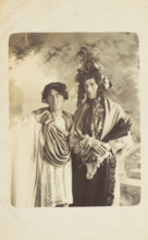 Two men in drag, one wearing a detailed feathered head garment, pose for a portrait, [1900-1909?]. Creator: Frank Wood.