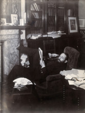 Arthur Schuster, lying on an armchair by a fireplace, with a photograph of Lord Kelvin..., c1901. Creator: Unknown.