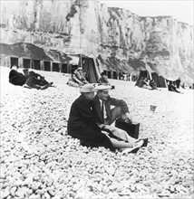 En 1938, à Etretat, couples assis sur une plage de galets au pied des falaises.