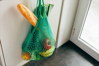 Green string shopping bag with vegetables, fruits and bread hanging on a hook in the kitchen