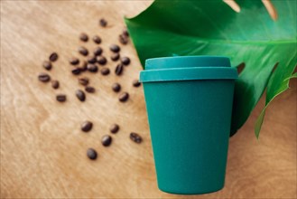 Zero waste concept, flat lay. Stylish reusable eco coffee cup  on wooden background with roasted coffee beans and green monstera leaf. Ban single use
