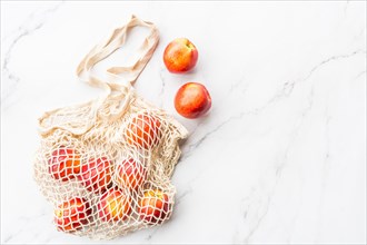 Fresh raw nectarines lying in string bag on white marble background. Flat lay, top view, summer, organic food, blogger style. Healthy concept