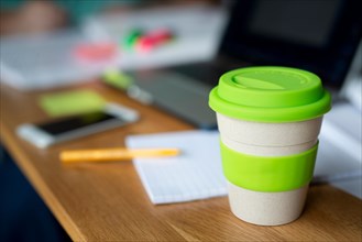 A bamboo reusable coffee cup sits on a wooden table along with other items such as text books and a laptop which suggests it's somebody's study space.