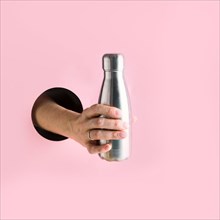 Metal reusable bottle in female hand through in pink hole. Trendy concept.