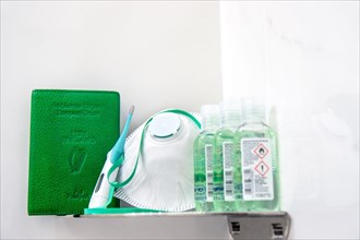 Coronavirus Covid-19 Ireland. Green Irish passport cover on shelf beside face mask respirator, hand sanitizer gel bottles and green thermometer stick