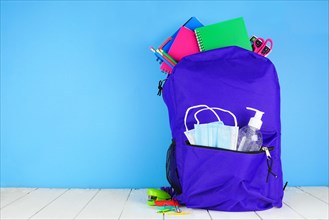 Backpack full of school supplies and COVID 19 prevention supplies. Blue background. Back to school during pandemic concept.