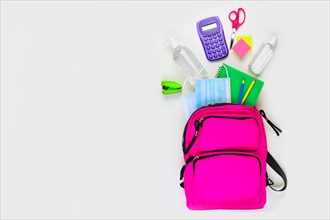 Backpack with school supplies and COVID 19 prevention items. Top view, spilling onto a white background. Back to school during pandemic concept.