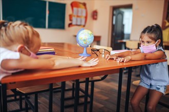 Two schoolgirls in medical masks are sitting at a school desk, opposite each other, group session, back to school, teaching children, social distance