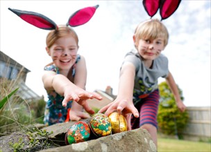 A brother & sister, wearing face paint & homemade bunny ears, take part in an Easter egg hunt on Easter Sunday in their garden.