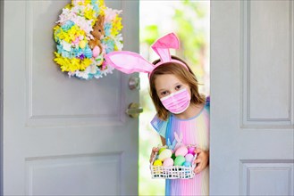 Little girl in face mask having fun on Easter egg hunt in covid-19 outbreak. Kids in bunny ears and rabbit costume in coronavirus pandemic celebration