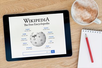 The Wikipedia website features on an iPad tablet device which rests on a wooden table beside a notepad and pencil and a cup of coffee (Editorial use o