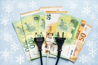 Electric plug lies on euro banknotes showing the drastical inflation on electricity costs at winter time. Energy crisis concept in Europe. Top view. Blue background with snowflakes