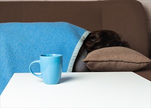 Blue Monday concept. There is a blue mug on the table, in the background is a girl under a blue blanket. Her posture demonstrates that she is upset or