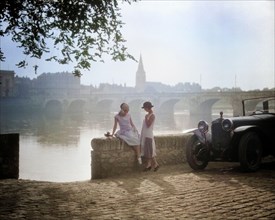 1920s TWO YOUNG WOMEN WITH ANTIQUE AUTOMOBILE SITTING STANDING TOGETHER TALKING BY STONE WALL LOIRE RIVER