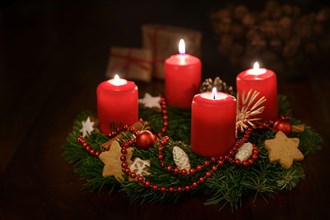 Advent wreath from fir branches with red lit candles and Christmas decoration, some gifts blurred in the dark background, copy space, selected focus,
