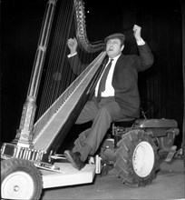 Comedian Raymond Devos rides a tractor playing harp
