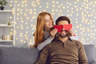 Woman covering boyfriend's eyes with red heart-shaped cards on Saint Valentine's Day