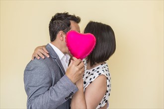 Side view of unrecognizable elegant couple kissing and hiding faces behind heart shaped balloon while expressing love on Saint Valentine Day on beige