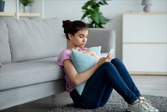 Stressed Indian teen girl sitting on floor with cellphone and reading bad message at home