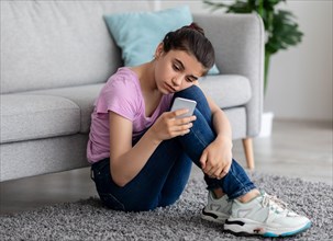 Unhappy Indian teen girl with mobile phone feeling sad, suffering from cyber bullying online, sitting on floor at home