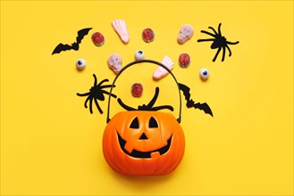 Happy Halloween. Top view of Halloween pumpkin basket full of candies, bats and spiders over yellow background. Halloween concept background